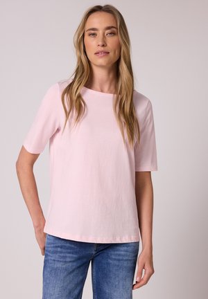 Femme aux longs cheveux blonds portant un haut à manches courtes rose clair et un jean bleu, debout les mains dans les poches devant un fond uni.