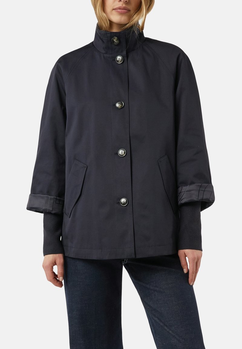 Frau trägt eine dunkelblaue, mit Knöpfen versehene Jacke mit Stehkragen und hochgekrempelten Ärmeln über einem schwarzen Langarmshirt und dunklen Jeans.