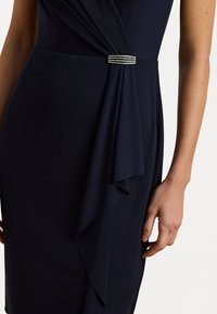 Námořnické šaty se drapovaným designem, s nařaseným korzetem a pasem zdobeným krystaly. Hladká tkanina s elegantním povrchem.