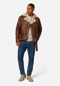 Brun läderjacka med fårskinnskrage, dragkedjefickor och belagd midja. Bärs över en ljus skjorta och blå jeans, kompletterad med beige stövlar.