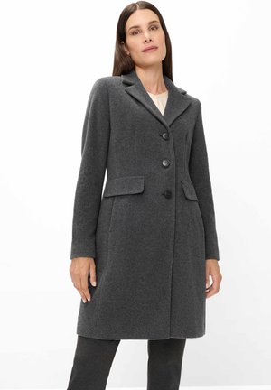 Frau trägt einen maßgeschneiderten, dunkelgrauen Mantel mit Knöpfen und Klappentaschen, steht vor einem weißen Hintergrund.