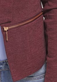 Giacca zip a maroon con tessuto testurizzato e zip dorata, dotata di tasca laterale e taglio sartoriale per un aspetto aderente.