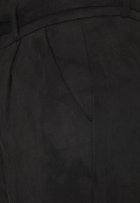 Pantalon noir sur mesure avec taille haute et plis, montrant une poche avant et une texture de tissu lisse.