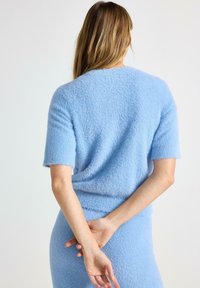Maglione azzurro chiaro morbido con maniche corte, caratterizzato da una texture soffice. Indossato con le mani incrociate dietro la schiena.