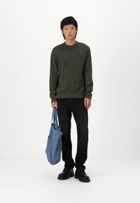 Mørkegrønn genser, svarte jeans og svarte sko. Holder en blå totebag. Enkel design med rund hals og ribbestrikkede mansjetter.