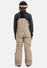 Personne portant un pantalon de neige beige et une veste noire avec un bonnet noir, se tenant de dos devant un fond uni.
