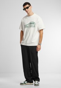 Tricou alb din bumbac cu text verde, asortat cu pantaloni negri largi și adidași verzi și albi. Bărbat purtând ochelari de soare dreptunghiulari, de culoare închisă.