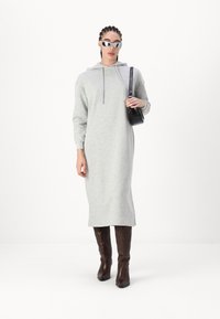Siva haljina dukserica s kapuljačom, dugim rukavima i bočnim prorezima, u kombinaciji s smešnim čizmama do kolena sa reljefnim uzorkom i malom crnom torbicom.