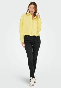 Gelbes, kurz geschnittenes Button-Down-Hemd mit einer einzelnen Tasche und langen Ärmeln, kombiniert mit schwarzen Skinny-Jeans und weißen Sneakers mit schwarzen Akzenten.
