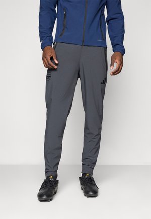 Grå treningsbukser med elastiske mansjetter, med Adidas-logo og lommepreg. Parret med svarte fotballsko og en marineblå jakke.