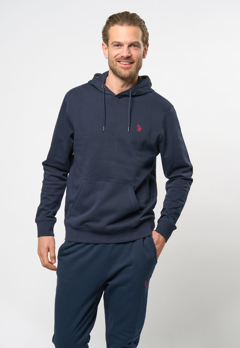 Marineblå hettegenser laget av mykt materiale med en frontlomme, justerbar snøring på hetta, og en liten rød logo på brystet. Kombinert med matchende bukser.