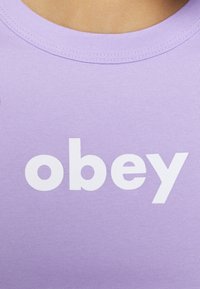 Fialové bavlněné tričko s kulatým výstřihem a výrazným bílým textem "obey" v moderním sans-serif písmu. Hladký povrch.