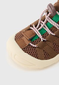 Καφέ πλεκτό αθλητικό παπούτσι με πράσινο λουράκι, κορδόνια λευκά με κόκκινες πιτσιλιές, δερμάτινες λεπτομέρειες και χοντρή λαστιχένια σόλα σε εκρού χρώμα, εμφανές σε λευκό φόντο.