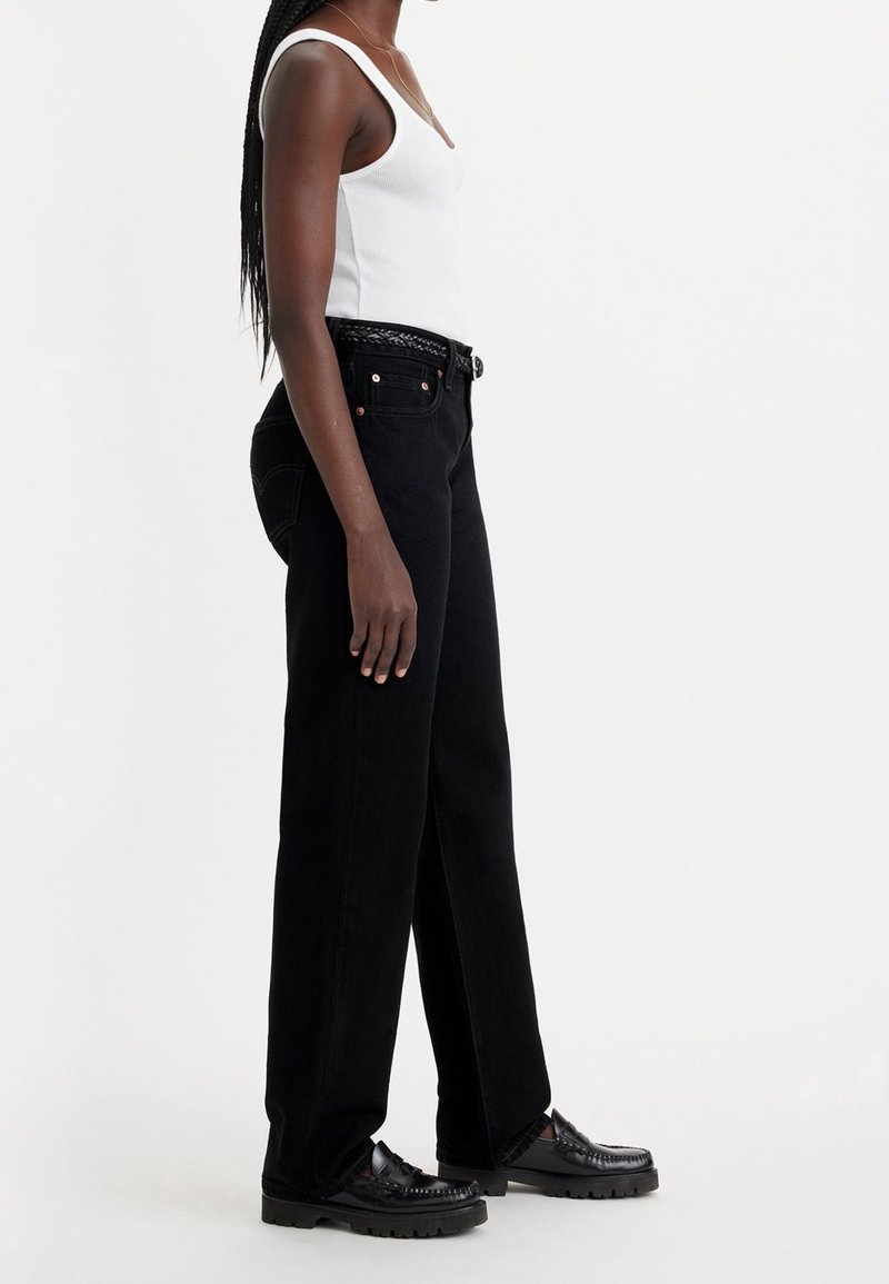 Femme portant un débardeur blanc, un jean noir taille haute à jambes larges avec une ceinture tissée, et des mocassins noirs épais, posant de profil.