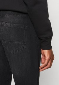 Persona vistiendo jeans de mezclilla negros y una camiseta de manga larga negra, con la mano derecha relajada a su lado contra un fondo blanco.