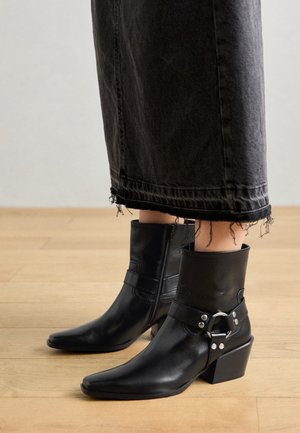 Personne portant des bottines noires à brides ornées de clous argentés et un jean noir raccourci aux bords effilochés.