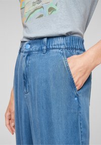 Close-up of light blue elastic-waist jeans with a hand in the right pocket and part of a gray graphic t-shirt visible.