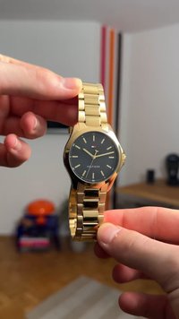 Goldfarbene Armbanduhr mit schwarzem Zifferblatt, ausgestattet mit einem runden Gehäuse, Stundenmarkierungen und einem Metallgliedband. Logo auf dem Zifferblatt sichtbar.