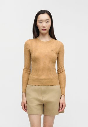 Donna con capelli lunghi e scuri che indossa un maglione aderente a coste color cammello e pantaloncini beige, in piedi davanti a uno sfondo chiaro e uniforme.