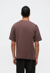 Muž stojaci chrbtom, oblečený v obyčajnom hnedom tričku s krátkym rukávom a čiernych nohaviciach na jednom svetlom pozadí.