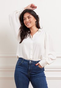 Blusa bianca con bottoni frontali, maniche a sbuffo e texture liscia, abbinata a jeans in denim blu scuro. Modella in posa con una mano alzata.