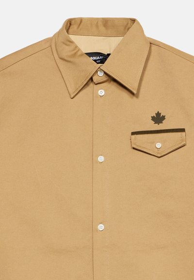 Chemise beige à boutons avec col pointu, poche poitrine avec bouton, et logo feuille d'érable brun foncé au-dessus de la poche.