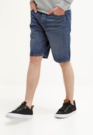 Mand iført blå denimshorts, grå langærmet trøje og sorte sneakers med hvide såler, stående med hænderne i lommerne.