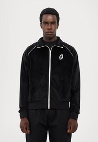 Veste en velours noir zippée avec des accents blancs et un logo. Elle dispose d'un col montant, de poignets et d'une taille côtelés. Les poches avant ne sont pas visibles.