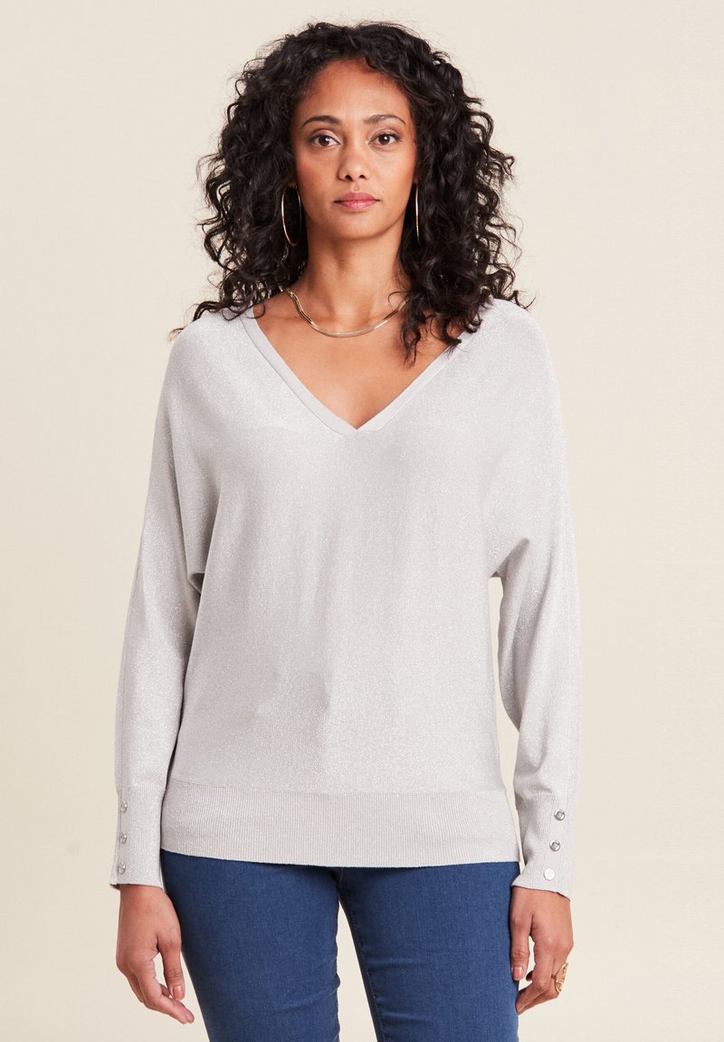 Silberner metallischer V-Ausschnitt-Pullover mit langen Ärmeln und Knopfakzenten an den Bündchen, kombiniert mit blauen Jeans. Weicher Stoff, lockere Passform.