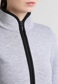 Giacca grigia con zip e colletto alto, caratterizzata da una zip nera e bordi neri. Tessuto morbido e testurizzato e design aderente nella parte superiore del corpo.