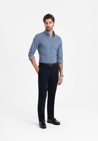 Camisa de botones azul claro con mangas remangadas, pantalones de cuadros oscuros, cinturón negro y zapatos negros, de pie contra un fondo blanco.