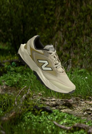 Chaussure de randonnée beige avec semelle robuste et logo « N », en l’air au-dessus d’un sol rocheux entouré de feuillage vert en milieu extérieur.