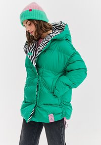 Žena oblečená v zelenej prešívanej bunde so zebrovým vzorom na podšívke, v ružovej a zelenej čiapke a čiernych džínsoch, stojí so zapichnutými rukami v vreckách.