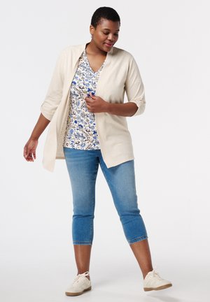 Beige vest over een bloemenprint top in blauw en wit, gecombineerd met cropped blauwe jeans en lichte sneakers. Soepele stof, casual pasvorm en opgerolde mouwen.