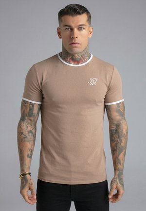 Young man with neck and arm tattoos wears beige fitted T-shirt with white trim and logo, standing against plain gray background.