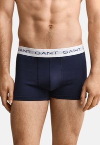 Male model wearing navy blue boxer briefs with white waistband branded "GANT" on a neutral background.