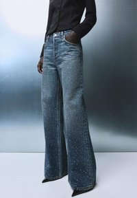Vaqueros de mezclilla azul con una cintura alta, piernas acampanadas y embellishments de strass dispersos. Cuenta con bolsillos y un cierre de botón.
