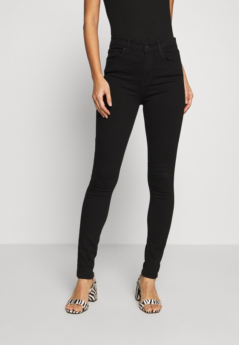 Femme portant un jean skinny noir taille haute et un top noir sans manches avec des sandales à talons ouvertes à motif zèbre sur un fond uni.