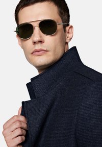 Niebieski, strukturalny płaszcz z wysokim kołnierzem, połączony z okrągłymi okularami przeciwsłonecznymi z ciemnymi soczewkami i złotymi oprawkami. Na tkaninie widoczne są subtelne detale.