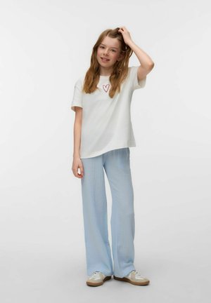 Dziewczyna o długich włosach, ubrana w białą koszulkę z czerwonym sercem, jasnoniebieskie spodnie i trampki, stoi z jedną ręką na głowie.