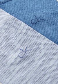 Calcetines rayados en azul claro y azul oscuro, con una textura de tela suave. Las iniciales de la marca "ck" están bordadas en un tono marino.