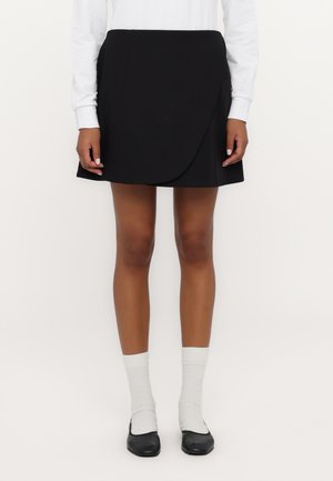 Zwarte wikkelrok van gladde stof, met een vooroverlapontwerp. Draagt een witte blouse met lange mouwen, witte sokken en zwarte ballerina's.