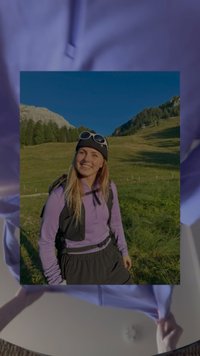 Lächelnde Frau in lilaner Jacke, schwarzem Hut und Sonnenbrille, die in einer grasbewachsenen Berglandschaft unter klarem blauen Himmel wandert.