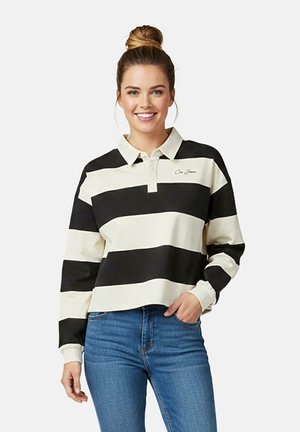 Jonge vrouw met haar in een knot, gekleed in een zwart-wit gestreept rugbyshirt met brede strepen en een blauwe spijkerbroek, glimlachend met één hand in haar zak.