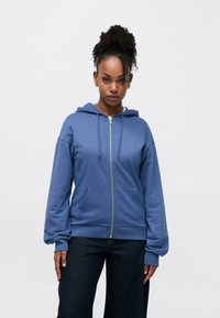 Μπλε φούτερ με φερμουάρ, φτιαγμένο από απαλό ύφασμα, με μπροστινή τσέπη, κουκούλα με κορδόνι και μανσέτες με ριμπ ύφανση, συνδυασμένο με σκοτεινό τζιν παντελόνι φαρδιάς γραμμής.