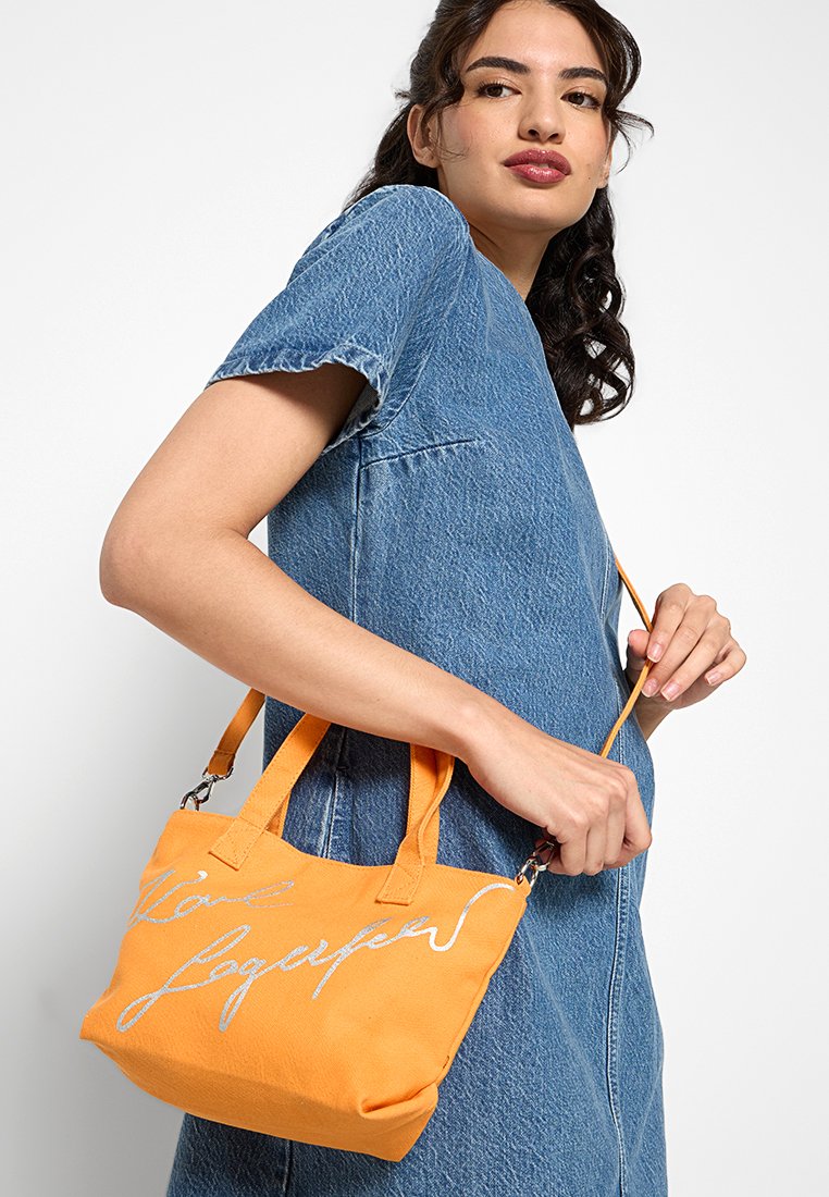 Bolso tote naranja con asas cortas, que presenta una inscripción en letra cursiva plateada. Hecho de material texturizado, tiene una forma estructurada y parte superior abierta.
