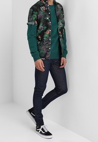 Černé tričko s krátkým rukávem a zeleným květinovým vzorem přes zelenou blůzu s dlouhým rukávem, v kombinaci s tmavě modrými džíny a černými teniskami.