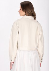 Crème leren cropped jack met een kraag, verlaagde schouders en knoopmanchetten. Gladde textuur, minimale stikseldetails.