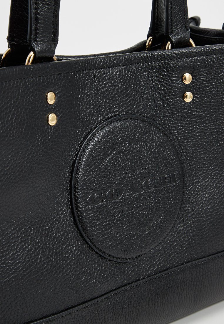 Bolso tote de cuero negro con superficie texturizada, acentos de hardware dorado y un parche circular con logotipo. Diseñado con asas dobles para llevar.