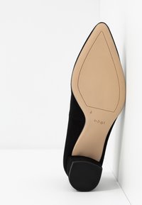 Escarpin en daim noir à talon aiguille avec un bout pointu, doté d'une semelle beige clair et d'une tige en tissu noir texturé. Logo de la marque sur la semelle.